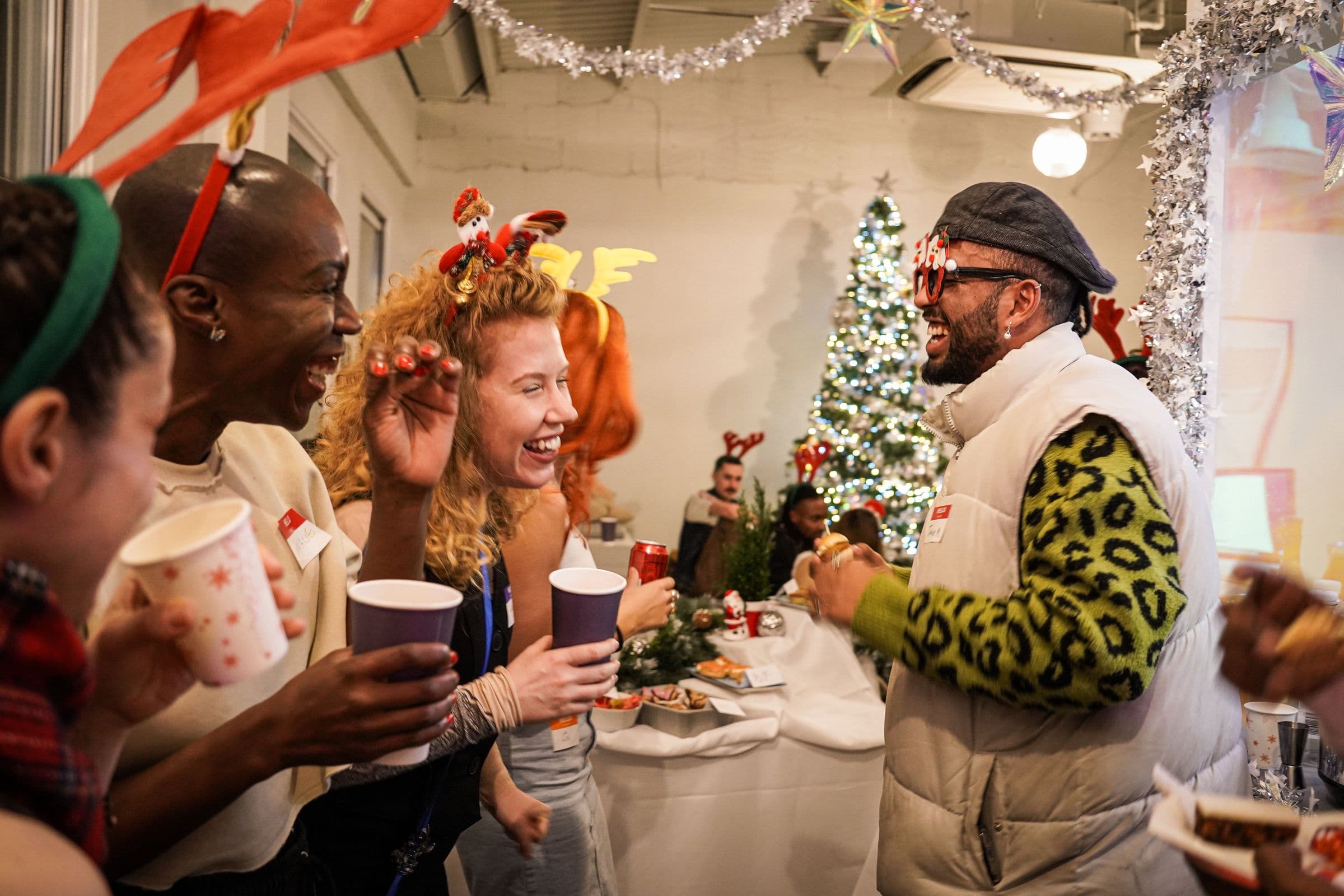 A group in reindeer antlers and festive glasses laughing over drinks beside a silver Christmas tree
