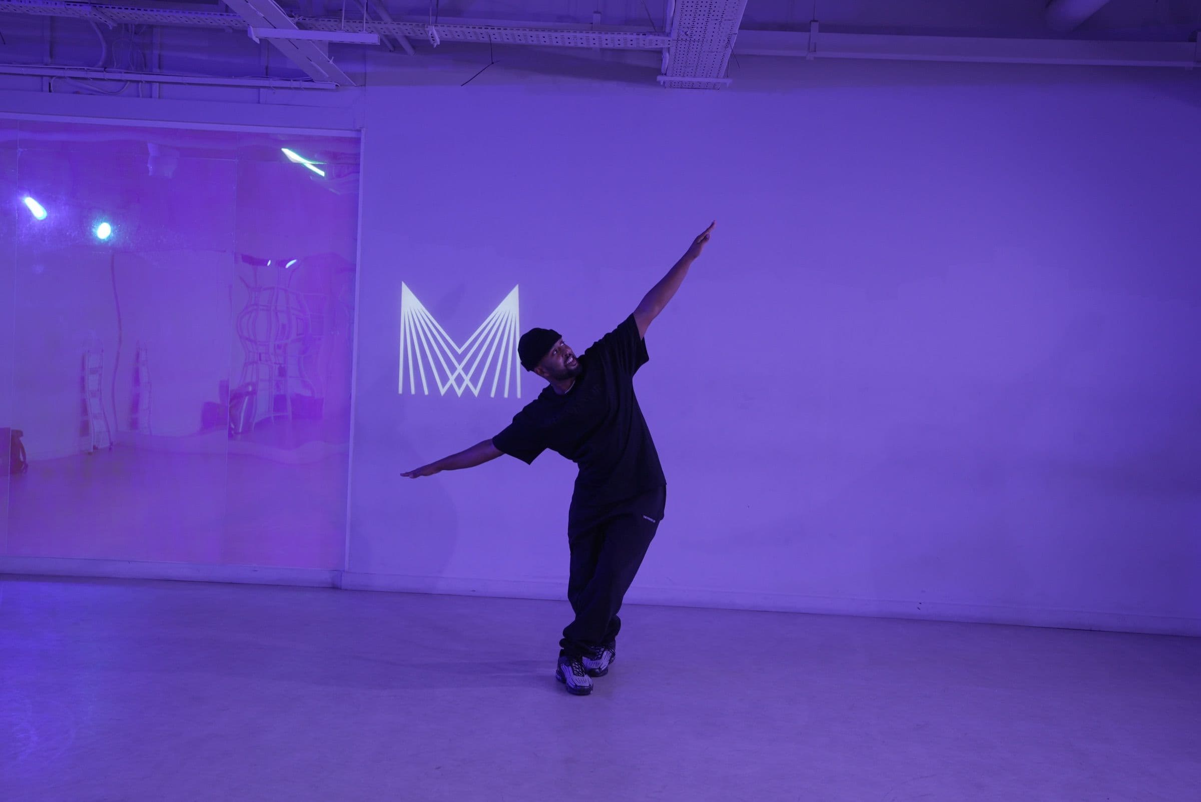 Jamie Mason mid-move in a Manor studio, one arm raised beside the projected Manor M, under purple light