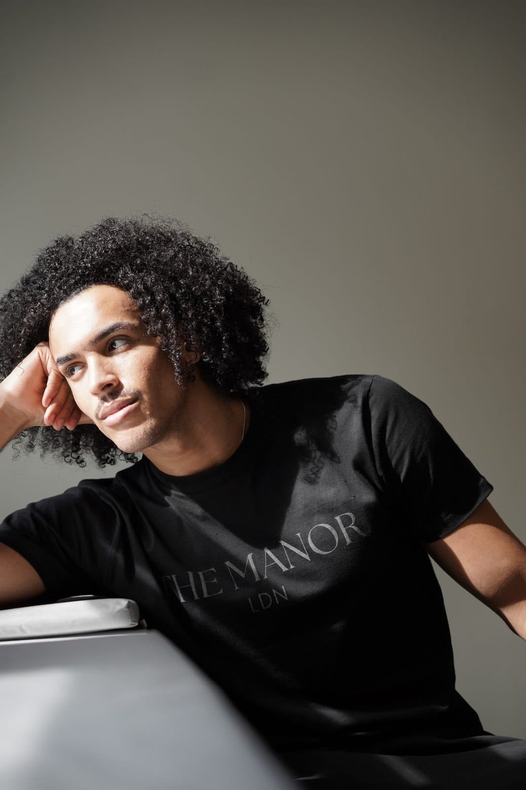 A dancer in the black THE MANOR LDN T-shirt, resting his head on his hand